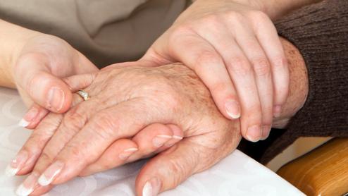 Zwei Hände, eine ältere und eine jüngere, liegen auf einem Tisch, wobei die jüngere Hand die ältere sanft umschließt.